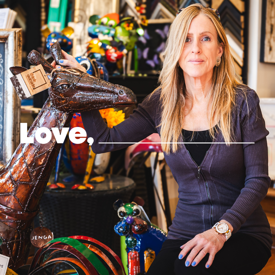 A woman with long blond hair and a purple sweater, kneels among colourfully painted metal sculptures in a shop. Her hand is on a sculpture of a giraffe.